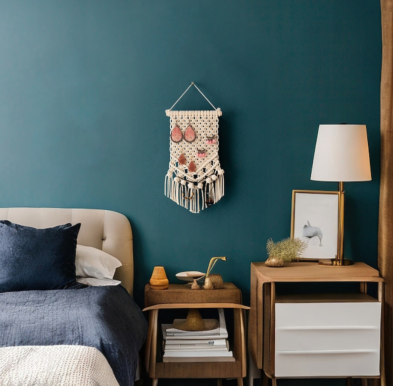 May include: A white macrame wall hanging with a geometric design and pink and white embellishments. The hanging is displayed on a teal wall above a wooden nightstand with a white lamp.