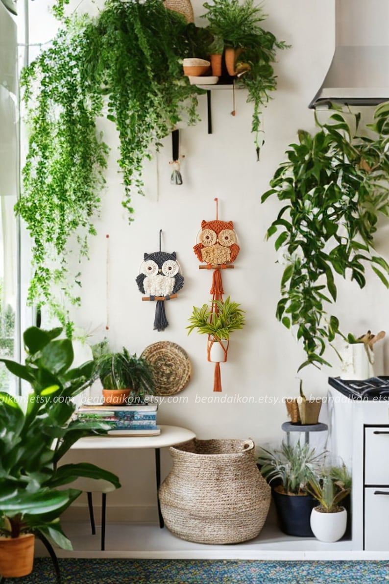 Peut inclure: Deux tentures murales en macram&eacute; en forme de hibou, l'une orange et l'autre grise, avec des accents de fil blanc. Les hiboux sont suspendus &agrave; un mur blanc avec des plantes en pots et un panier tiss&eacute; sur le sol.