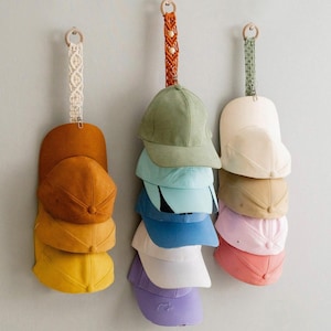 May include: Three hanging hat organisers with multiple baseball caps in various colours. Each organiser has a wooden ring and a decorative strap. The hats include shades of yellow, brown, blue, green, and pink.
