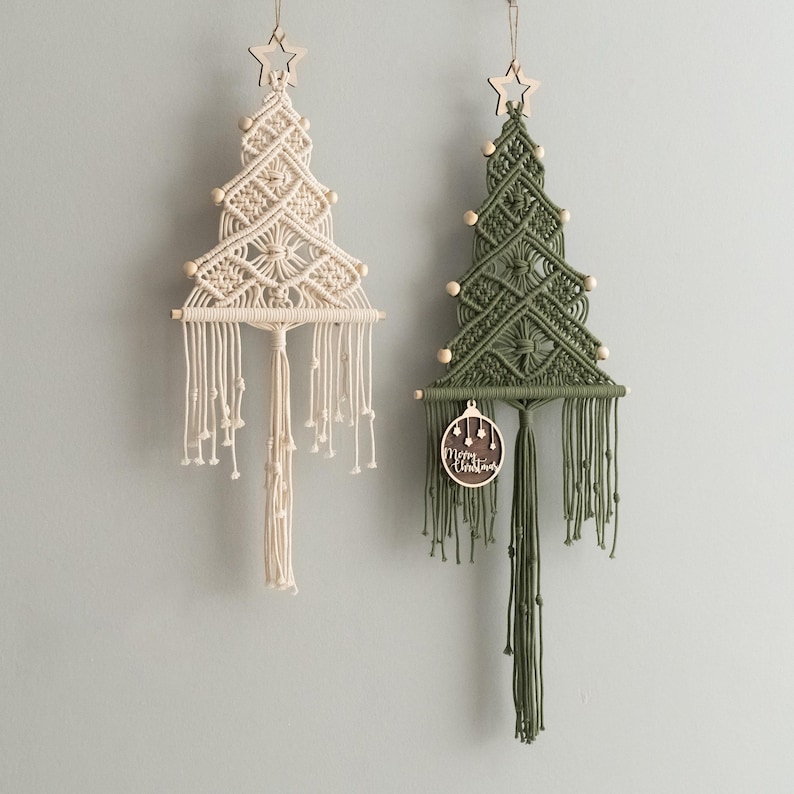May include: Two macrame wall hangings shaped like Christmas trees, one cream and one olive green. Each features a wooden star at the top, wooden beads, and a horizontal wooden dowel. The green tree includes a round wooden ornament with the text Merry Christmas.