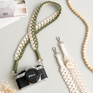 May include: Two camera straps, one green and white and one white, with silver clasps. The green and white strap is looped around a silver camera with a black lens. The white strap has a wooden tag with the name "Mia" on it.