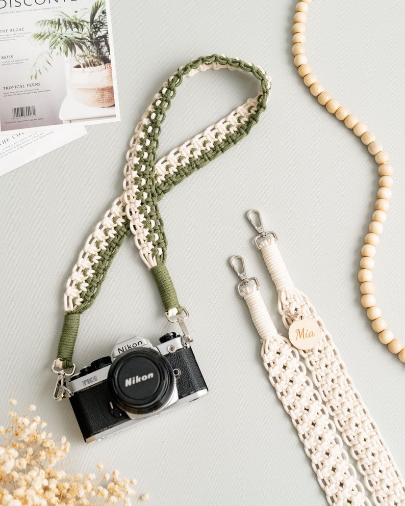 May include: Two camera straps, one green and white and one white, with silver clasps. The green and white strap is looped around a silver camera with a black lens. The white strap has a wooden tag with the name "Mia" on it.