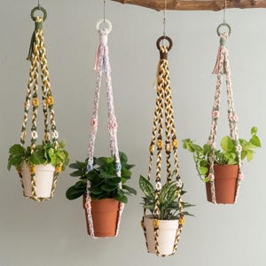 Può includere: Quattro portavasi in macramè di vari colori, ciascuno con una pianta in vaso. I portavasi sono appesi a un ramo. I vasi sono bianchi e terracotta. I disegni in macramè incorporano giallo, bianco, marrone e rosa.