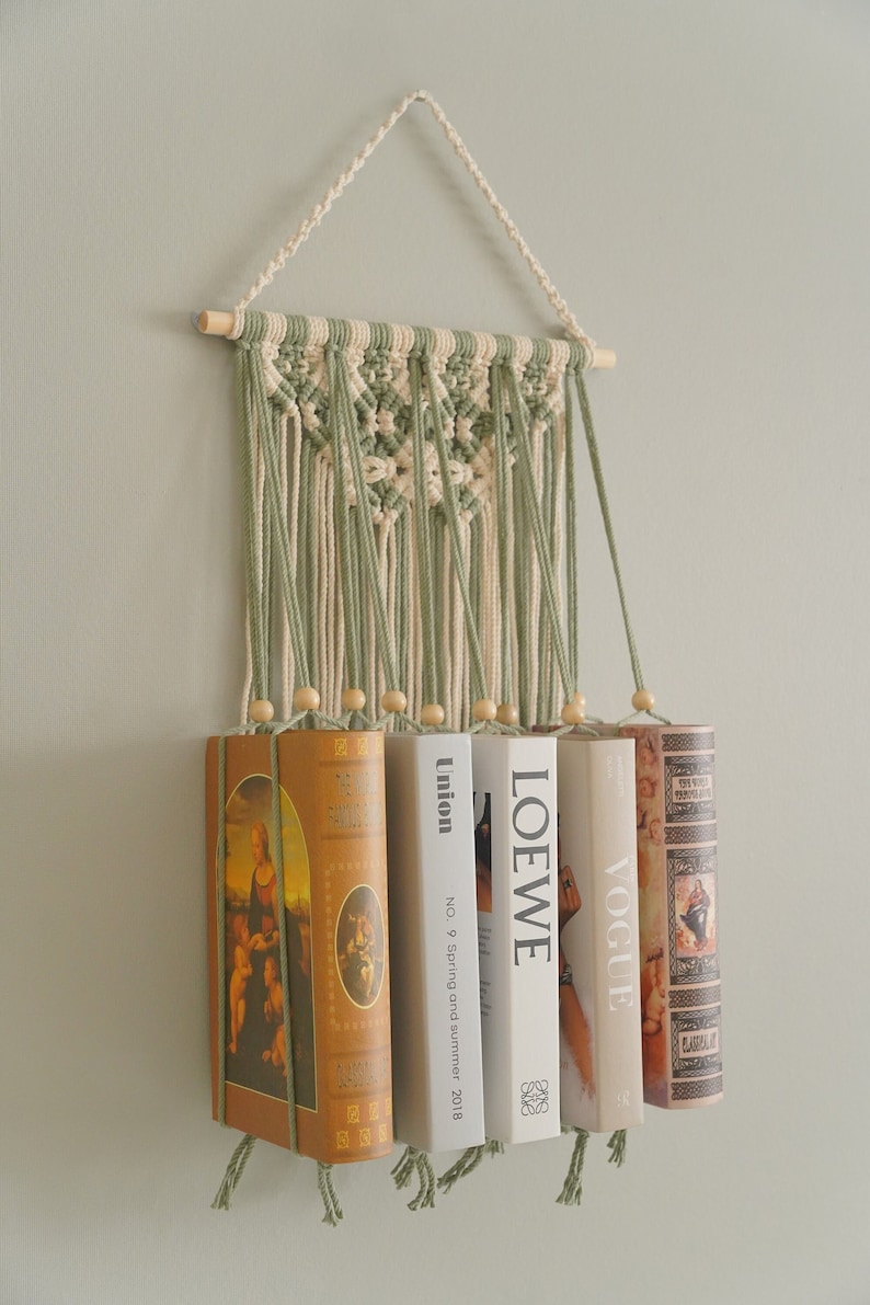K&ouml;nnte beinhalten: Ein Wandbehang aus Makramee mit einer Holzleiste und gr&uuml;nem und cremefarbenem Garn. Der Behang wird verwendet, um eine Reihe von B&uuml;chern zu halten, darunter Vogue, LOEWE, Union und ein Buch mit einem Gem&auml;lde der Madonna mit Kind.