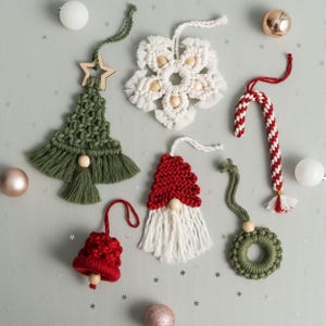 May include: A selection of handmade Christmas decorations in various shapes and colours. The ornaments include a green macrame tree, a white snowflake, a red and white candy cane, a red bell, a gnome, and a green wreath. Each decoration has a string for hanging.
