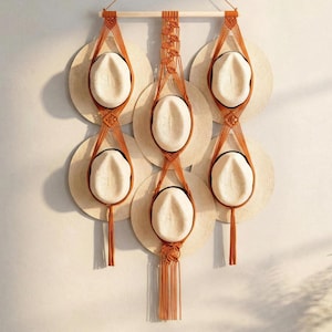 May include: A macramé hat rack in shades of brown, holding six straw hats with white crowns. The rack is suspended from a wooden dowel, with decorative knots and tassels. The hats are arranged in a visually appealing pattern.