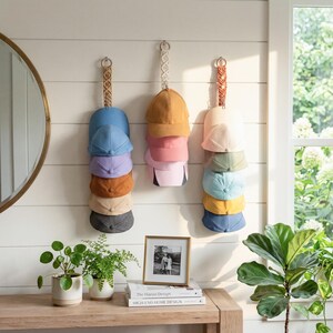 May include: A selection of baseball caps in different colours, displayed on a white wall. The caps are arranged in vertical stacks, featuring shades of blue, pink, yellow, and neutral tones. The display is part of a home decor setting.