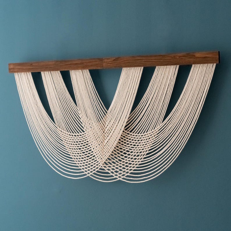 May include: A wall hanging featuring a horizontal dark brown wooden bar from which multiple strands of off-white cotton cord are suspended. The cords are arranged in four distinct, overlapping U-shaped swags that create a layered effect.