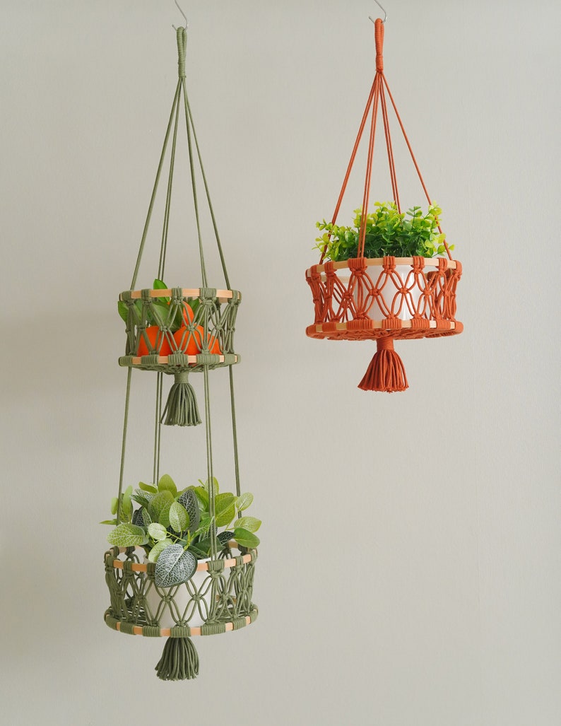 May include: Two macrame plant hangers with wooden rings. The top hanger is green and has two tiers, one with oranges and the other with a green plant. The bottom hanger is orange and has a single tier with a green plant.