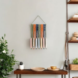 May include: A macrame wall hanging with a wooden dowel and a geometric pattern in shades of green, orange, and blue. The hanging is decorated with books.