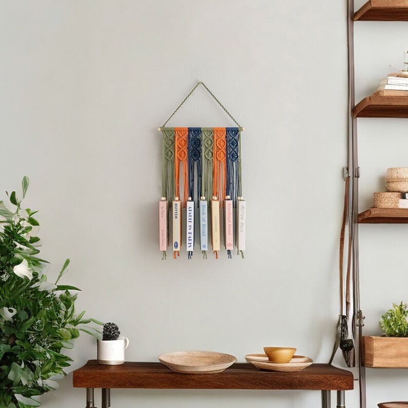 May include: A macrame wall hanging with a wooden dowel and a geometric pattern in shades of green, orange, and blue. The hanging is decorated with books.