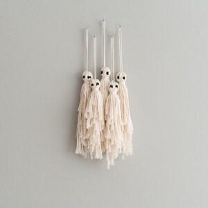 May include: Five white macrame tassels with black accents hanging from a wall. The tassels are made of a soft, fluffy material and have a simple, elegant design.
