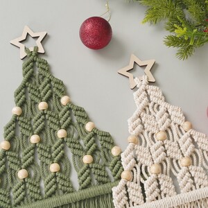 May include: Two macrame Christmas trees with wooden star ornaments and wooden beads. The trees are green and white and are hanging from a white wall.