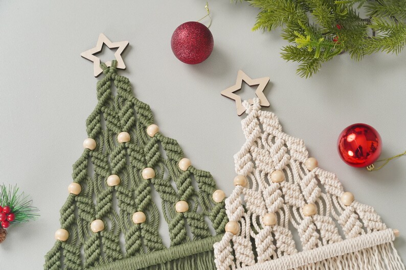 May include: Two macrame Christmas trees with wooden star ornaments and wooden beads. The trees are green and white and are hanging from a white wall.