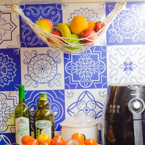 May include: A kitchen counter with a white and blue patterned tile backsplash. A white hammock with fruit hanging from it is attached to the top of the backsplash. A basket of red tomatoes sits on the counter in front of two bottles of oil and a black air fryer. The air fryer has the word "PHILIPS" printed on it.