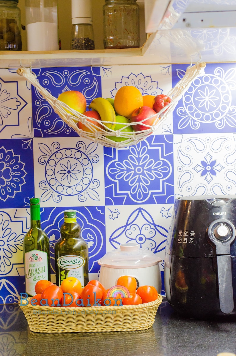 May include: A kitchen counter with a white and blue patterned tile backsplash. A white hammock with fruit hanging from it is attached to the top of the backsplash. A basket of red tomatoes sits on the counter in front of two bottles of oil and a black air fryer. The air fryer has the word "PHILIPS" printed on it.