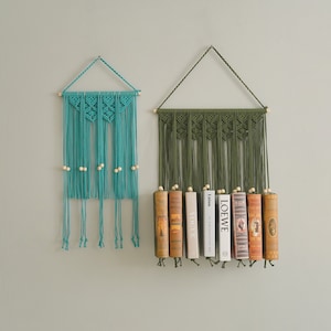 May include: Two macrame wall hangings with wooden beads. The top one is teal blue and the bottom one is green. The green one has books displayed on it.