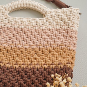May include: A crocheted bag with a brown strap and a multi-colored design. The bag is made of a thick, textured yarn and has a light brown, pink, and brown color scheme.