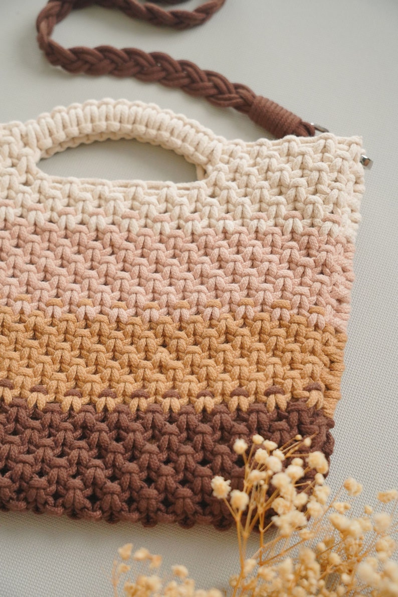 May include: A crocheted bag with a brown strap and a multi-colored design. The bag is made of a thick, textured yarn and has a light brown, pink, and brown color scheme.