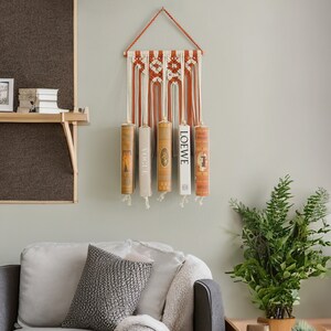 K&ouml;nnte beinhalten: Ein Wandbehang aus Makramee mit einem geometrischen Muster in Orange und Wei&szlig;. F&uuml;nf B&uuml;cher werden an dem Behang ausgestellt, wobei der Titel "LOEWE" auf einem der B&uuml;cher sichtbar ist.