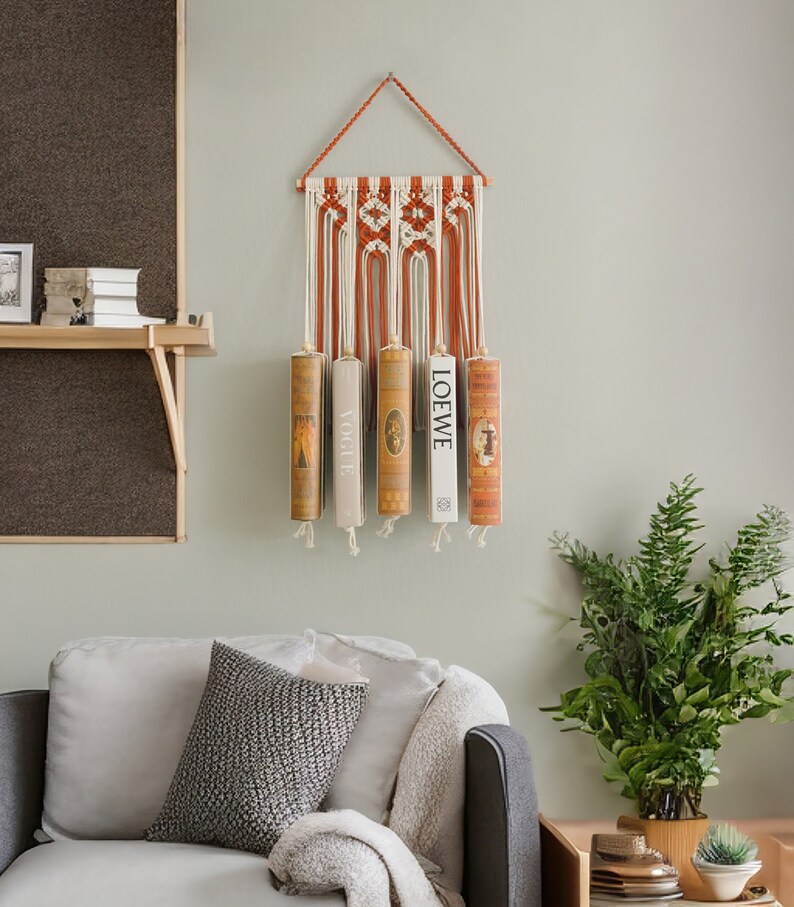 K&ouml;nnte beinhalten: Ein Wandbehang aus Makramee mit einem geometrischen Muster in Orange und Wei&szlig;. F&uuml;nf B&uuml;cher werden an dem Behang ausgestellt, wobei der Titel "LOEWE" auf einem der B&uuml;cher sichtbar ist.