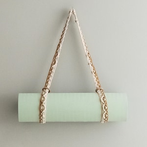 May include: A light green yoga mat hanging on a wall with a white and brown macrame strap.