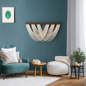 May include: A wall hanging featuring a dark wooden horizontal bar with off-white cotton cord draped in multiple overlapping U-shaped loops. The cord is attached to the top bar and hangs down in a symmetrical, layered pattern across the wall.