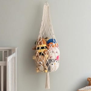 May include: A cream-colored macrame hanging storage basket filled with plush toys. The basket is suspended from a wall hook and features a diamond-patterned net design. The toys include a bear, a bee, a cat, and a penguin.