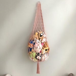 May include: A pink macrame hanging storage basket filled with various stuffed animals. The basket is suspended from a wall and features a woven net design with a tassel at the bottom. The stuffed animals are of different colors and sizes.