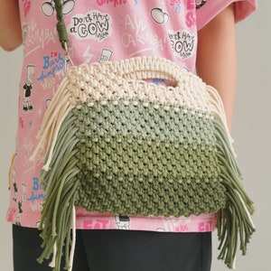 May include: A green, white, and beige macrame bag with long fringe. The bag is worn over the shoulder of a person wearing a pink t-shirt with a cartoon print.