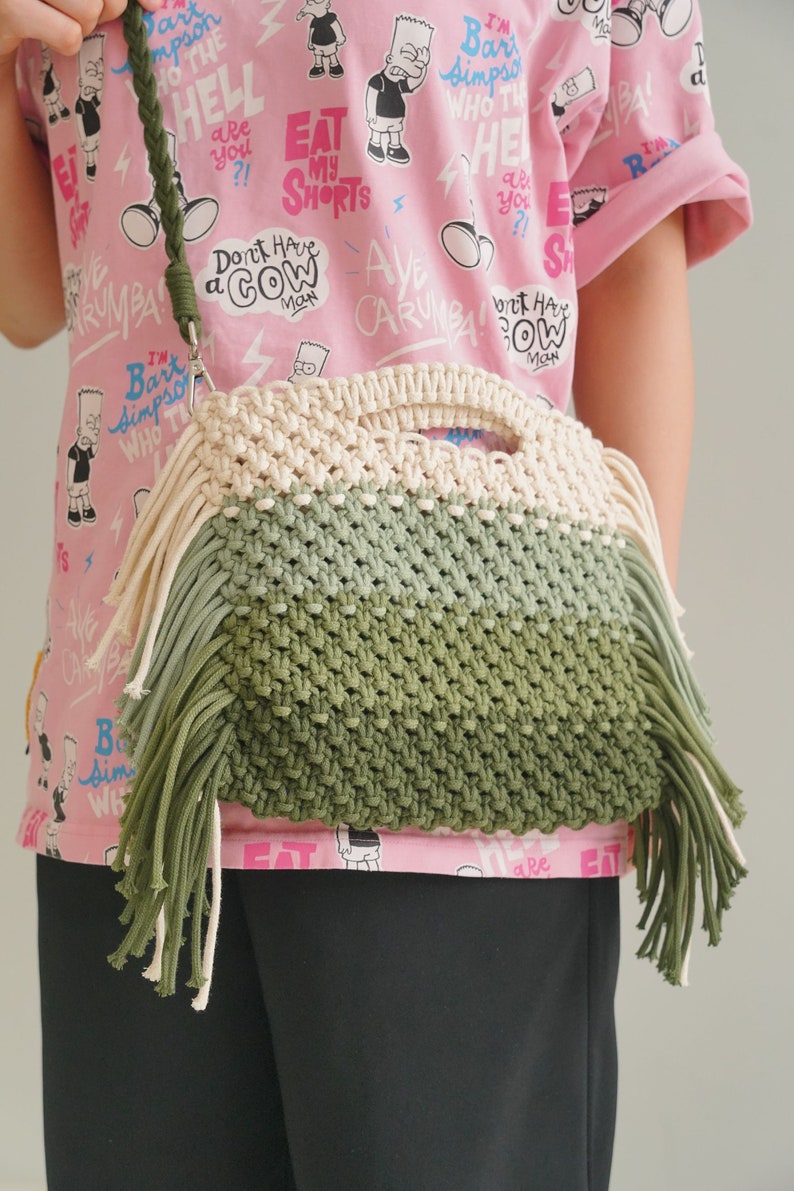 May include: A green, white, and beige macrame bag with long fringe. The bag is worn over the shoulder of a person wearing a pink t-shirt with a cartoon print.