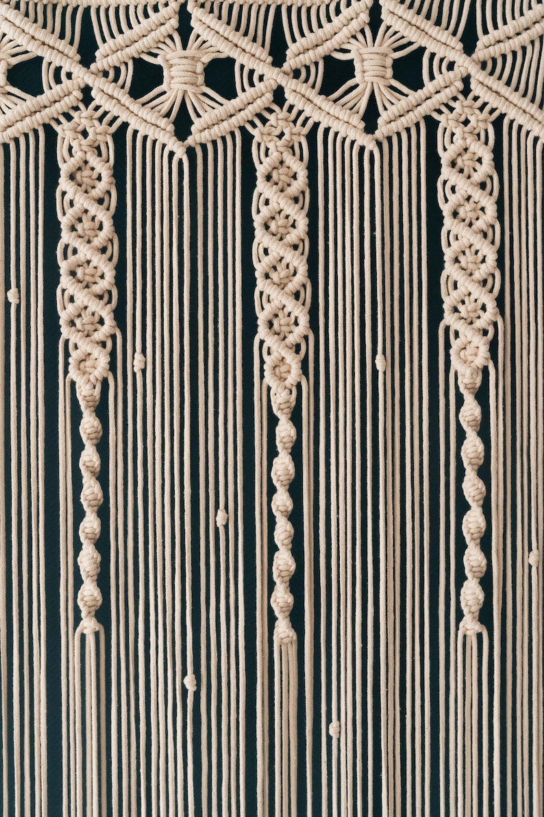 K&ouml;nnte beinhalten: Ein beigefarbener Makramee-Wandbehang mit geometrischem Muster. Der Behang besteht aus gekn&uuml;pften Schn&uuml;ren und hat unten einen Fransen.