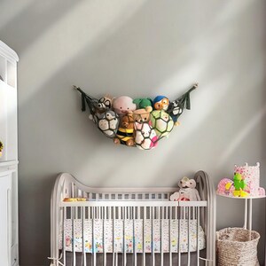 Op de afbeelding: Een wit wiegje met een wit en bloemig beddengoed. Een groene en zwarte net hangmat gevuld met knuffels hangt aan de muur boven de wieg. Een kleine tafel met een rieten mand en een roze en groene knuffel staat in de hoek van de kamer.