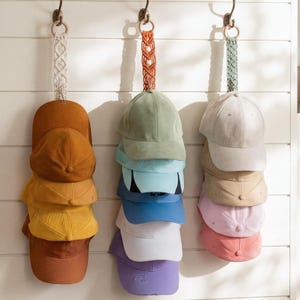 Peut inclure: Trois ensembles de casquettes de baseball suspendues à un mur blanc. Chaque ensemble est suspendu par une sangle macramé décorative fixée à un crochet en métal. Les casquettes sont de différentes couleurs, dont le marron, le vert, le bleu et le rose.