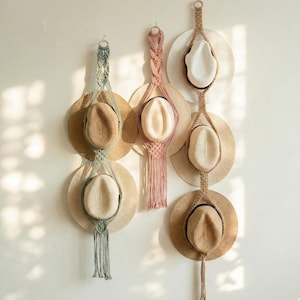 May include: Macrame hat holders in beige, pink, and green, each holding a straw hat. The hats are displayed against a white wall, creating a bohemian aesthetic. The holders are made of woven rope.