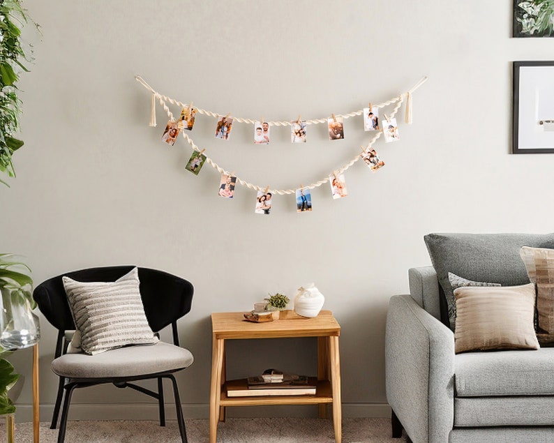 Peut inclure: Une guirlande de photos en corde blanche avec des glands est accroch&eacute;e &agrave; un mur blanc. La corde est enfil&eacute;e de petites photos de personnes, et des pinces &agrave; linge en bois maintiennent les photos en place. Un canap&eacute; gris avec des coussins se trouve au premier plan.