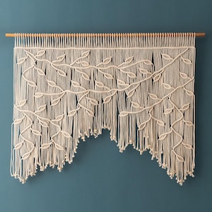 Pode incluir: Uma tapeçaria de macramé branca com um padrão de folhas. A tapeçaria é feita de cordão de algodão e tem uma vareta de madeira na parte superior.