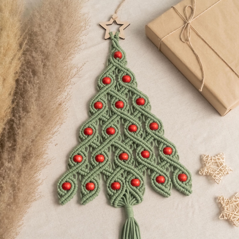 K&ouml;nnte beinhalten: Ein gr&uuml;ner Makramee-Weihnachtsbaum-Wandbehang mit roten Holzperlen. Der Baum ist mit einem Holzstern an der Spitze verziert und hat eine Quaste am Boden.