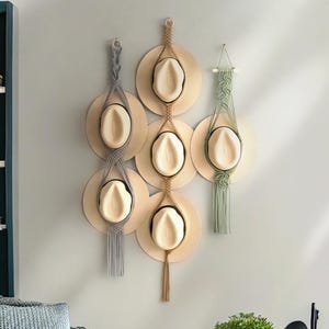 Op de afbeelding: Een verzameling strohoeden die aan een muur worden getoond met behulp van macramé-hangers. De hoeden zijn beige met zwarte banden, en de hangers zijn in grijstinten, beige en groen. De hoeden zijn in een decoratief patroon gerangschikt.