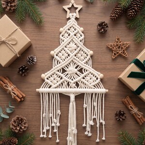 May include: A cream-coloured macrame wall hanging shaped like a Christmas tree, featuring intricate knotted patterns, wooden beads, and a wooden star topper. The design includes a horizontal wooden dowel at the base with long, knotted fringe tassels hanging down.