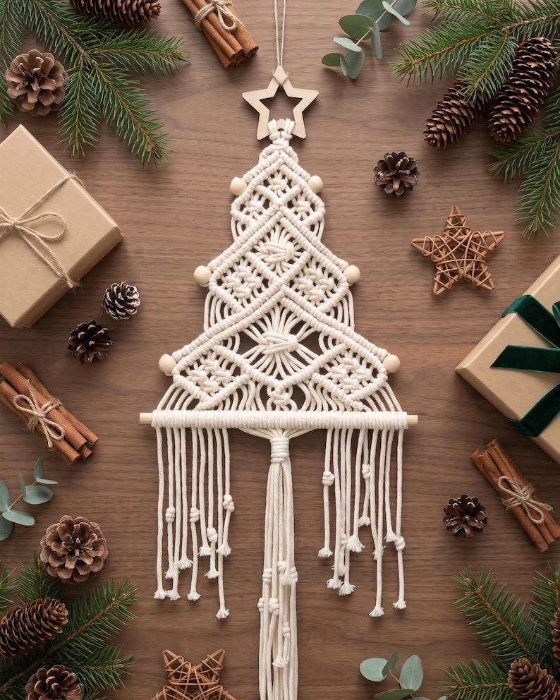 May include: A cream-coloured macrame wall hanging shaped like a Christmas tree, featuring intricate knotted patterns, wooden beads, and a wooden star topper. The design includes a horizontal wooden dowel at the base with long, knotted fringe tassels hanging down.