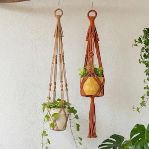 Peut inclure: Deux suspensions macramé, une beige et une couleur rouille, chacune contenant une plante en pot. Le support beige a un pot blanc, tandis que celui de couleur rouille a un pot jaune. Les deux suspensions sont suspendues à une branche.