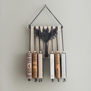 May include: A macrame wall hanging with a geometric design in black and cream, holding six vintage books. The books are suspended by cords with wooden beads. The wall hanging is mounted on a light gray wall.