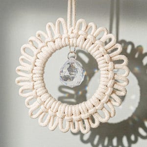 May include: A cream-colored macrame wreath with a clear crystal prism hanging in the center. The wreath is circular with a looped design. The crystal prism is faceted, reflecting light. A cream-colored cord is attached for hanging.