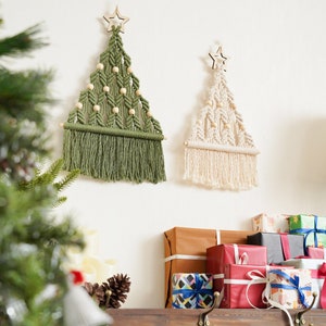 Puede incluir: Dos &aacute;rboles de Navidad de macram&eacute; con estrellas de madera en la parte superior, uno verde y otro crema, colgados en una pared blanca sobre un estante de madera con regalos envueltos.