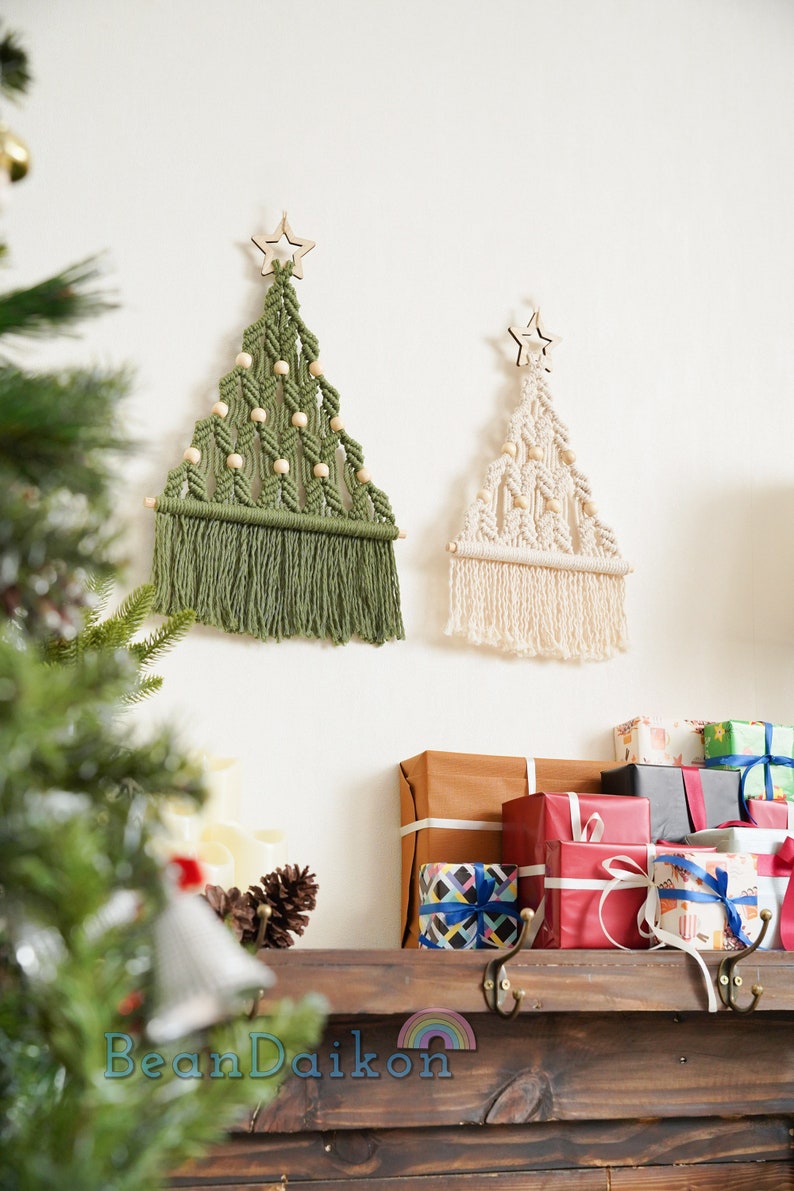 Puede incluir: Dos &aacute;rboles de Navidad de macram&eacute; con estrellas de madera en la parte superior, uno verde y otro crema, colgados en una pared blanca sobre un estante de madera con regalos envueltos.