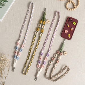 以下が含まれることがあります： ピンク、ブルー、グリーン、パープルなど、さまざまな色の編み込み携帯電話ストラップのコレクション。各ストラップは花の模様と金属製の留め金が付いています。1つは、チャームで飾られたマルーンの携帯電話ケースに取り付けられています。