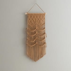 May include: A beige macrame wall hanging with a geometric pattern and fringe. The hanging is made of natural fibers and has a rustic, bohemian style.