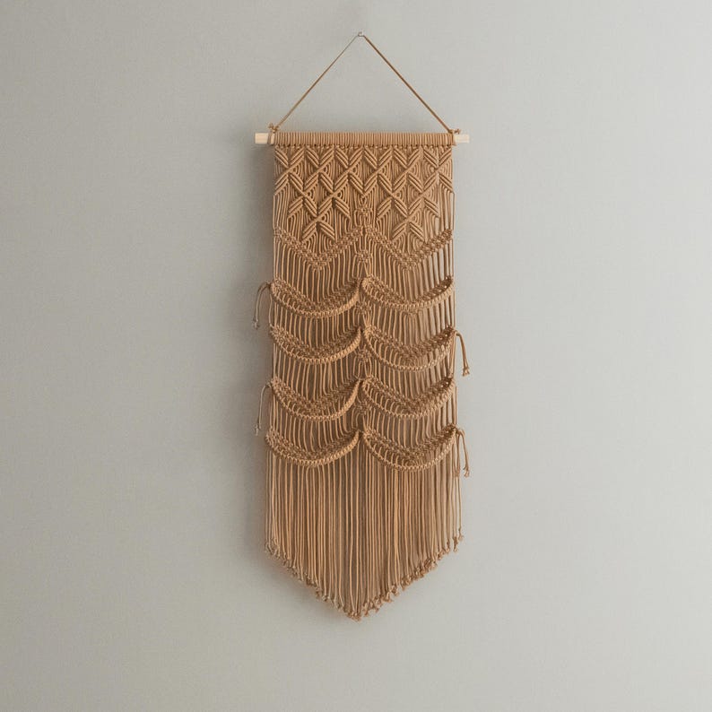 May include: A beige macrame wall hanging with a geometric pattern and fringe. The hanging is made of natural fibers and has a rustic, bohemian style.