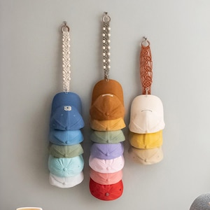 Puede incluir: Una pared con tres colgadores de pared de macramé, cada uno sosteniendo una pila de gorras de béisbol. Las gorras son de varios colores, incluyendo azul, amarillo, verde, morado, rosa y rojo.